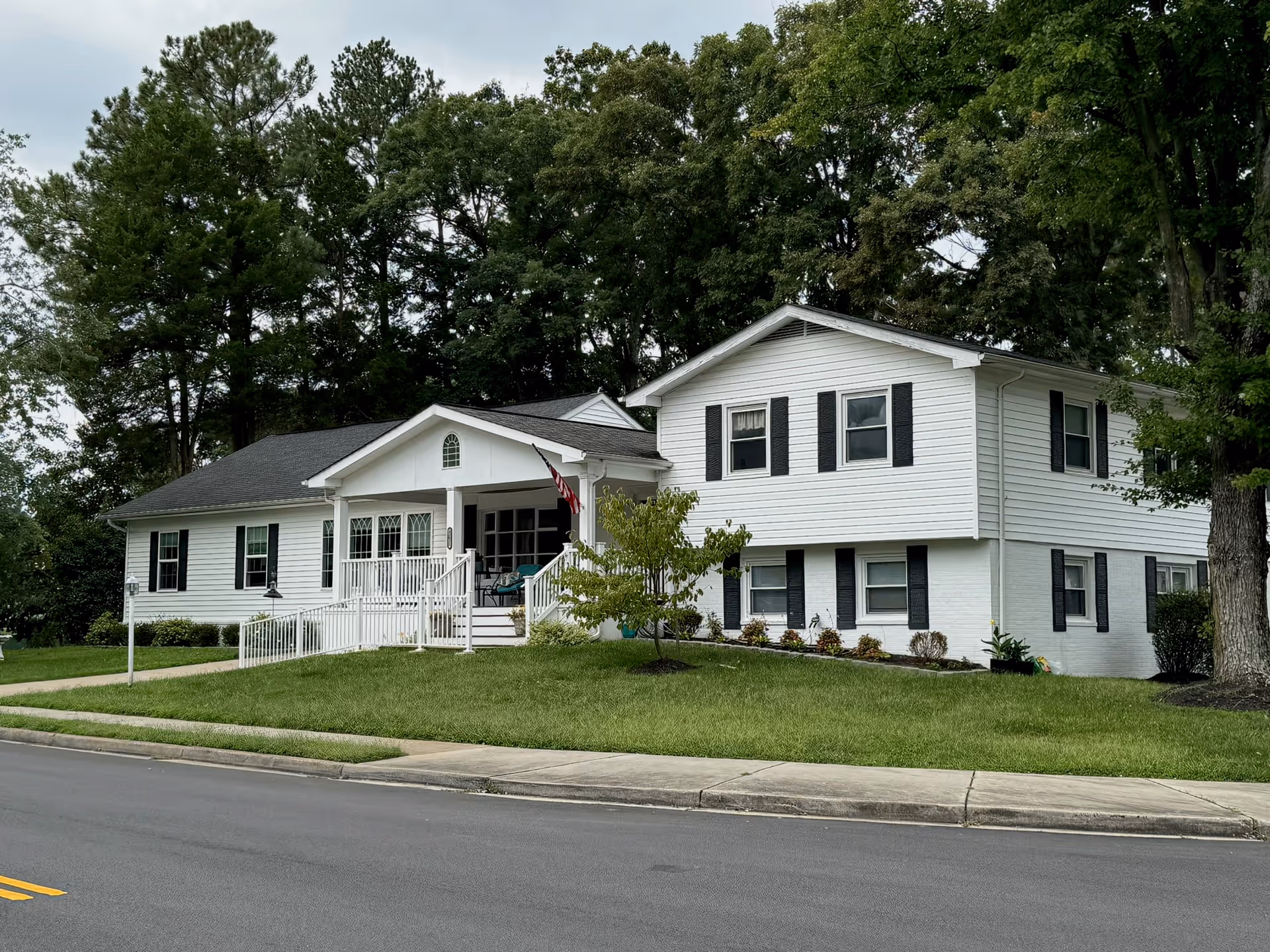 Exterior view of a white two-story assisted living facility building with black shutters, a front porch with a white railing and steps, a small tree and landscaped bushes in the front yard, and a paved road in the foreground. Tall trees are visible in the background.