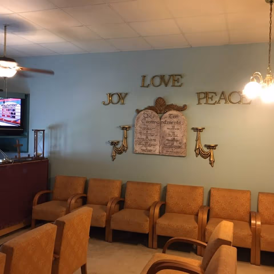A room with a row of six orange upholstered chairs with wooden armrests against a light blue wall. On the wall are decorative gold letters spelling 'JOY', 'LOVE', and 'PEACE' around a plaque displaying the Ten Commandments. There are also two gold wall-mounted candle holders on either side of the plaque. A ceiling fan and a hanging light fixture are visible, along with a TV mounted on the left side showing a game or quiz program.