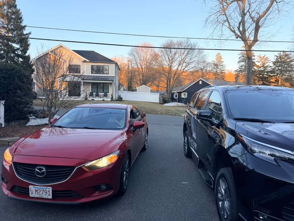 Two cars parked on a residential street with houses and trees in the background.