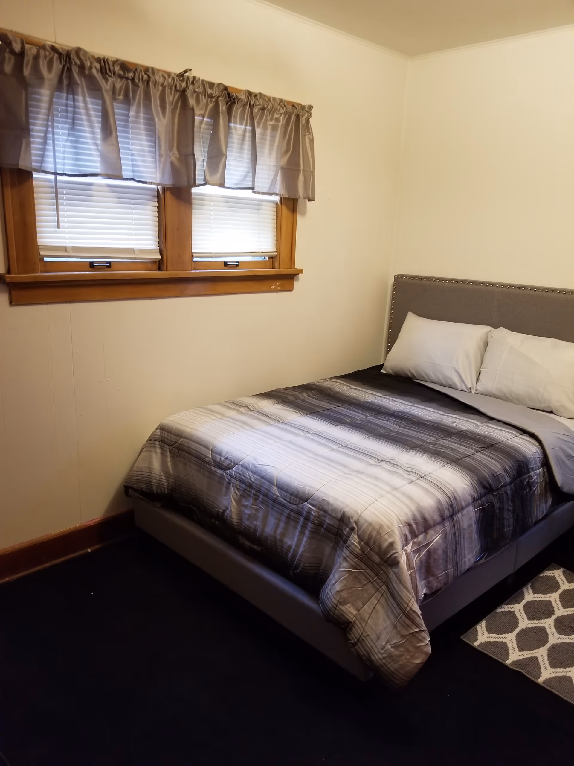 A furnished bedroom featuring a gray upholstered bed, windows, blinds, and a patterned rug.