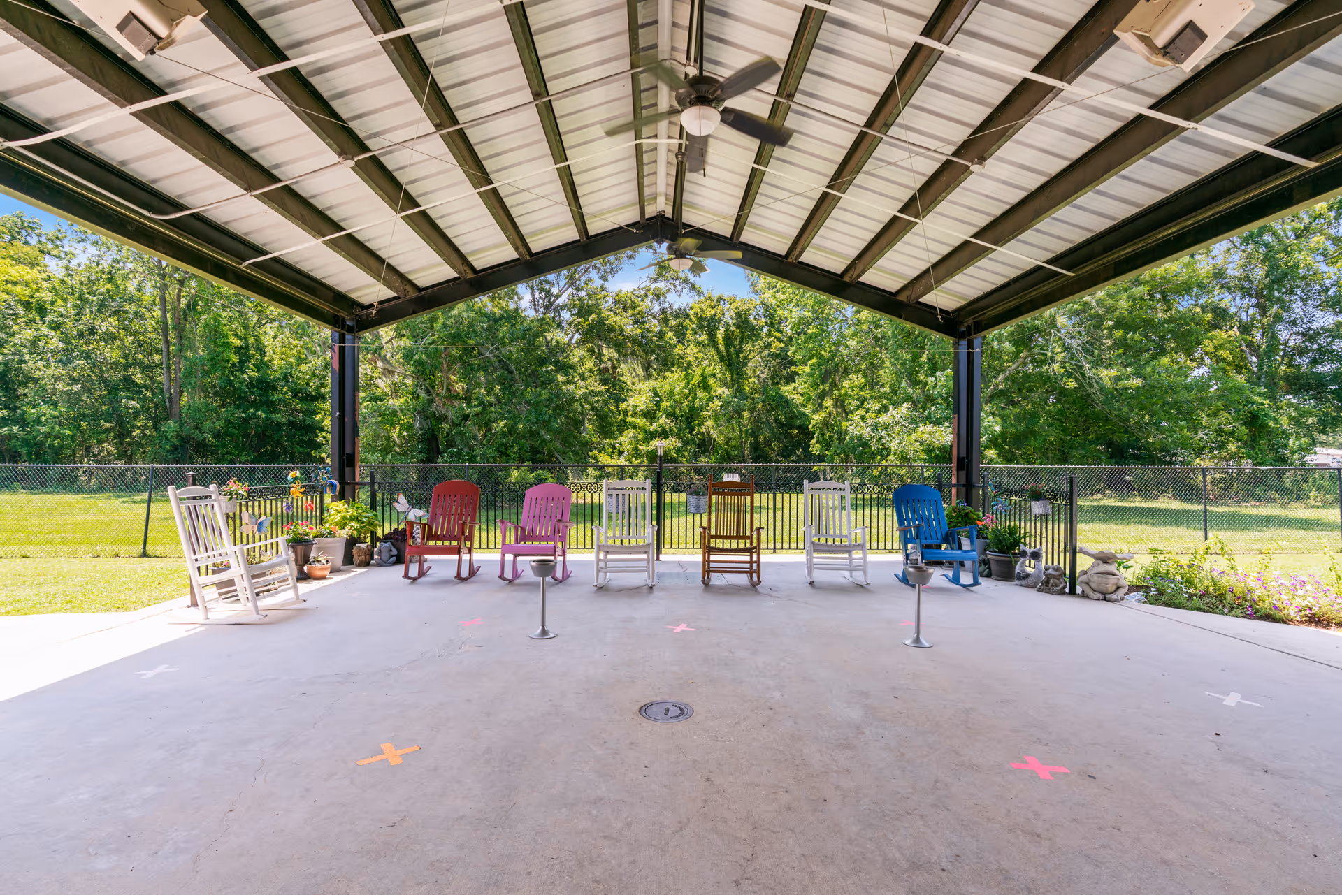 Covered outdoor patio area with a metal roof and several colorful rocking chairs arranged in a row facing outward. The patio overlooks a grassy area with trees and is surrounded by a black chain-link fence. There are potted plants placed along the fence.