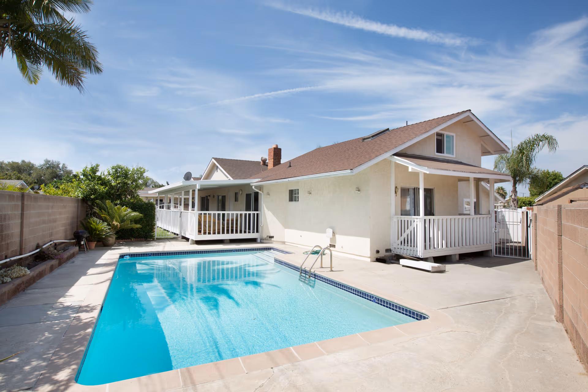 Outdoor view of a senior care facility showing a clear blue swimming pool surrounded by a concrete deck. The building has a beige exterior with a brown roof, white railings, and a covered porch area. There are palm trees and other greenery around the pool area under a partly cloudy blue sky.