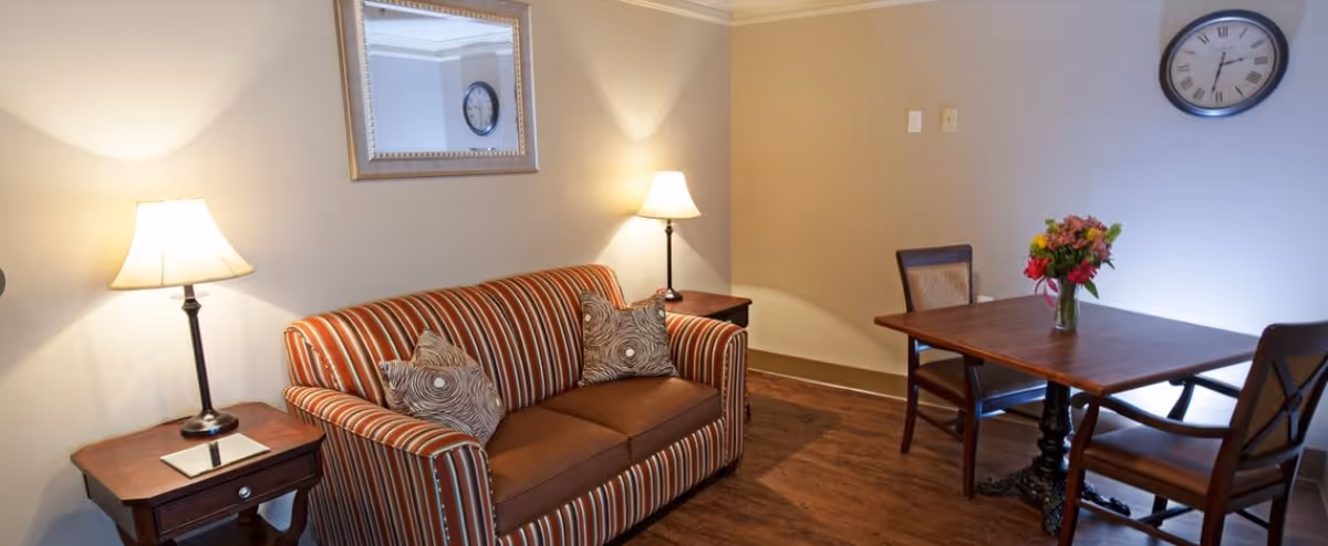 A cozy living room area with a striped sofa featuring two patterned cushions, two side tables each with a lamp, a wooden dining table with two chairs, a vase of flowers on the table, a wall clock, and a large mirror reflecting part of the room.