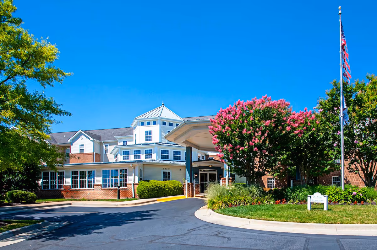 Entrance and front facade of a multi-story senior living building with a covered porte-cochere, flowering trees, and an American flag.