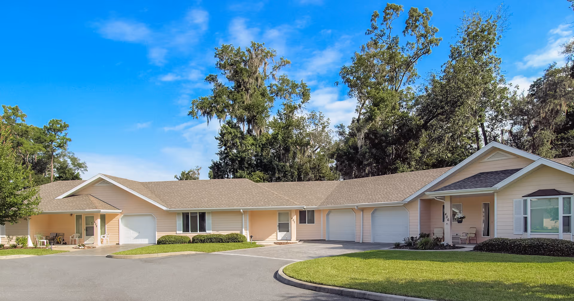 Exterior view of a single-story residential building with a light peach exterior and multiple garage doors, surrounded by green grass and tall trees under a blue sky with some clouds.