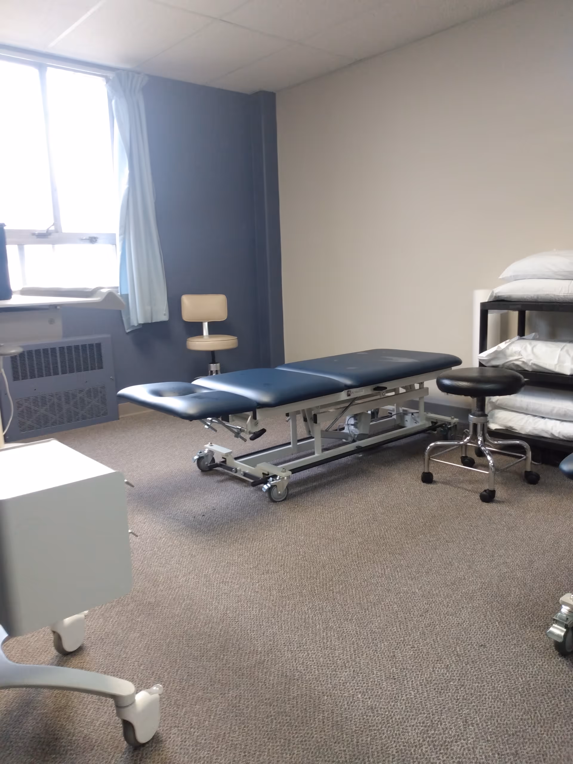 A medical examination room with a blue padded table, stools, and stacked pillows.