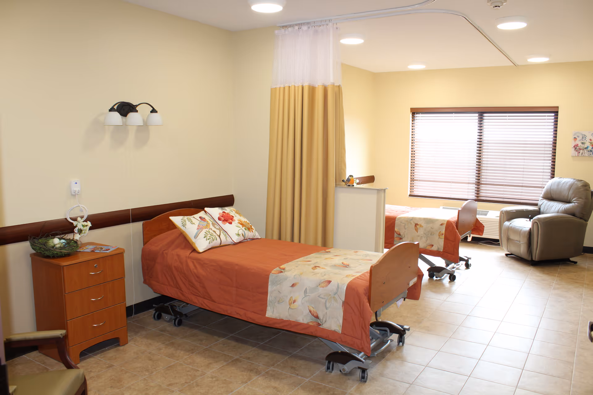 Two hospital-style beds with orange bedding and a recliner chair in a shared patient room.