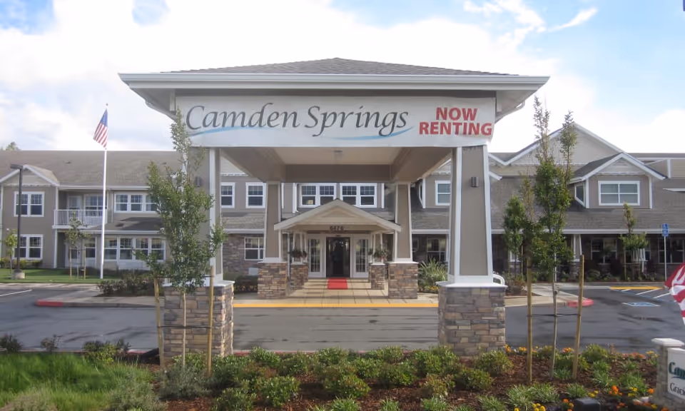 Front exterior view of Camden Springs Gracious Retirement Living facility with a covered entrance displaying a sign that reads 'Camden Springs NOW RENTING'. The building is two stories with multiple windows, surrounded by landscaped greenery and a parking area.