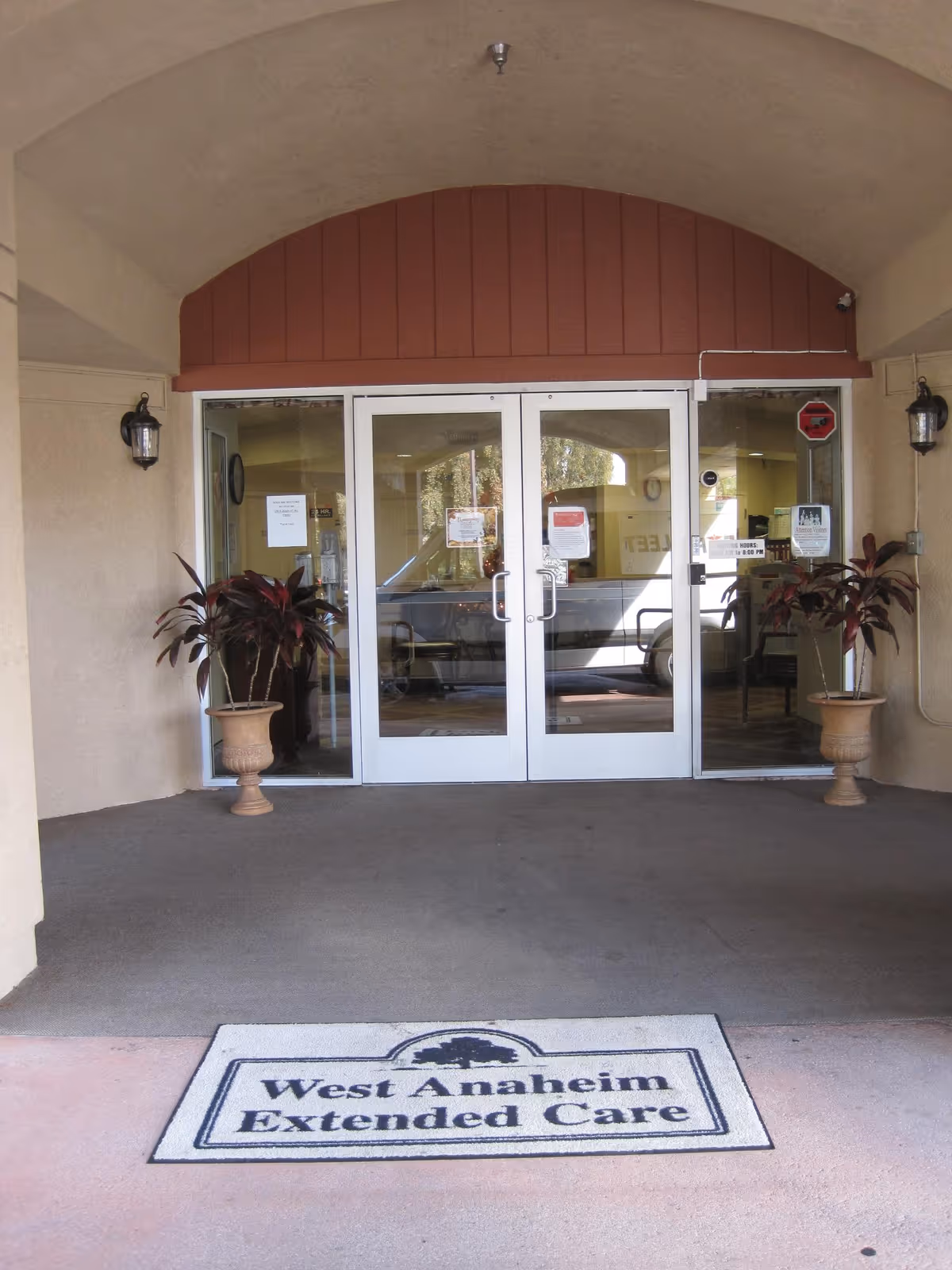 Entrance to a care facility with double glass doors, two potted plants on either side, and a doormat that reads 'West Anaheim Extended Care'. The entrance is under an arched overhang with beige walls and two wall-mounted lantern lights.