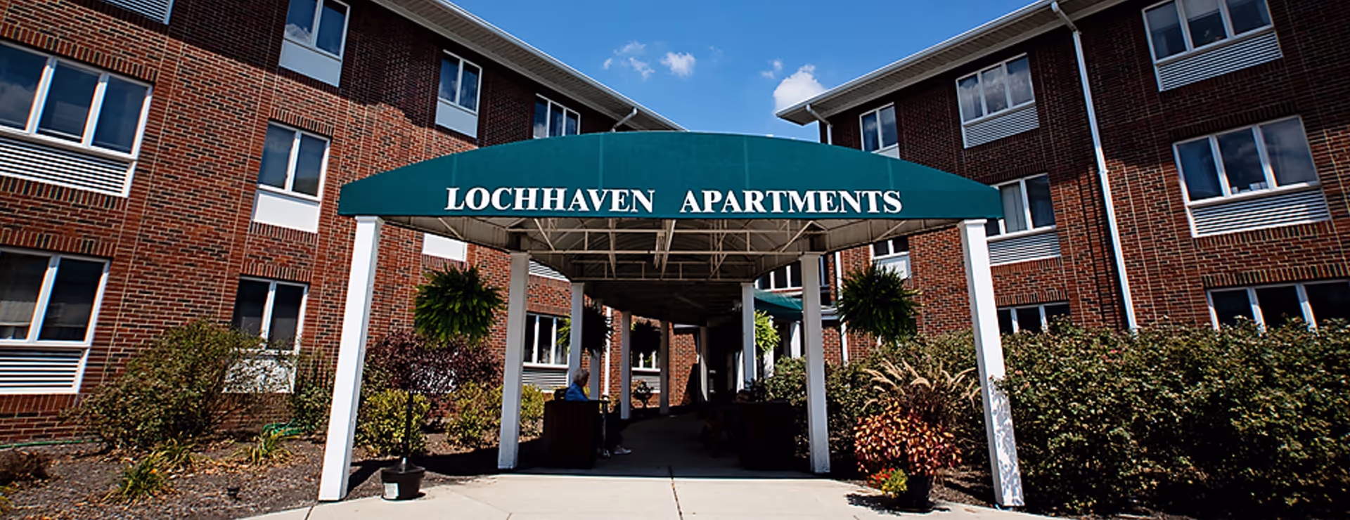 Entrance to Lochhaven Apartments with a green canopy supported by white pillars. The building is made of red brick with multiple windows. There are plants and shrubs on either side of the walkway leading to the entrance, and a person is sitting on a bench under the canopy.