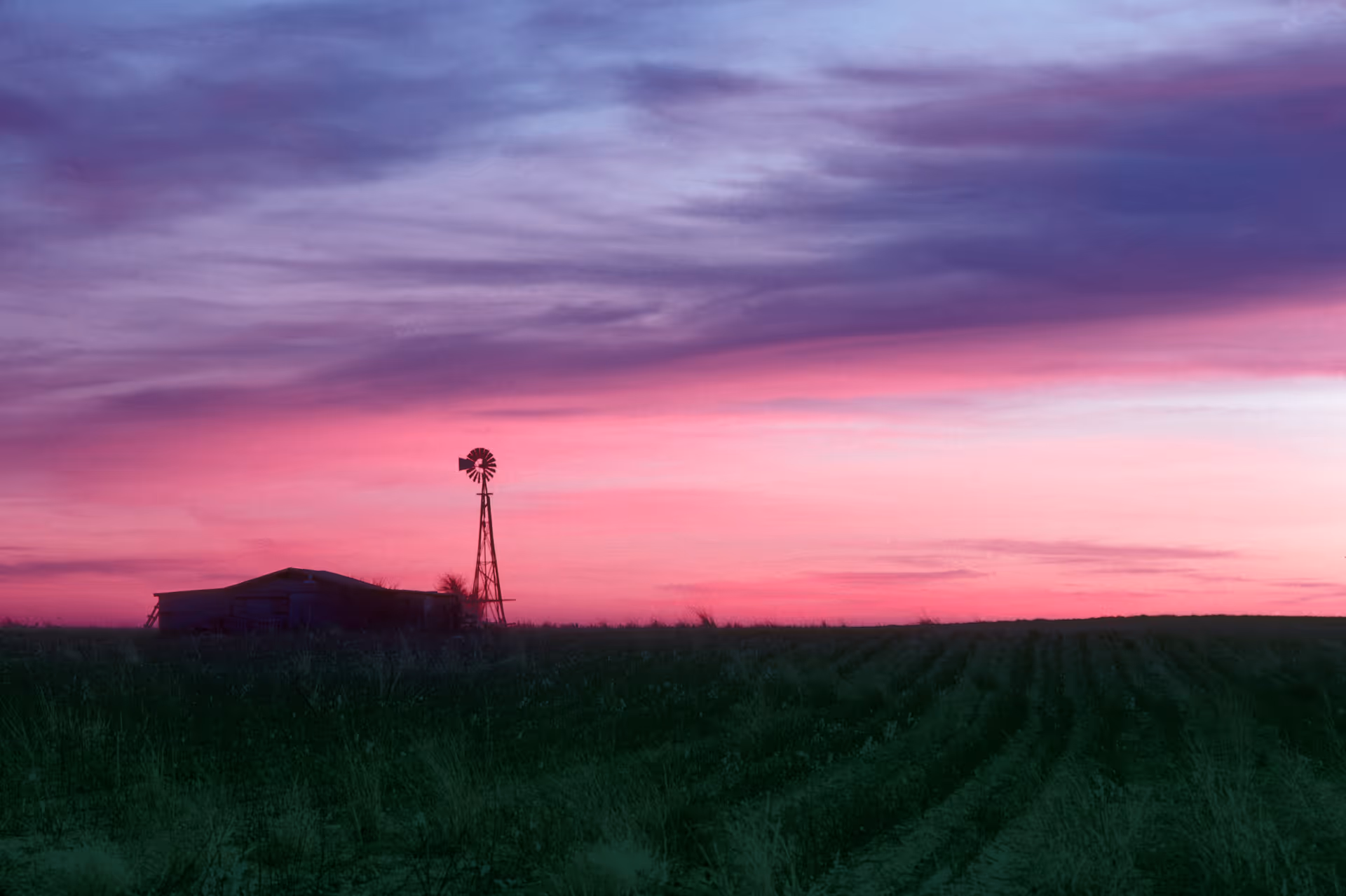 Sunset over a rural field with a windmill and barn silhouette.