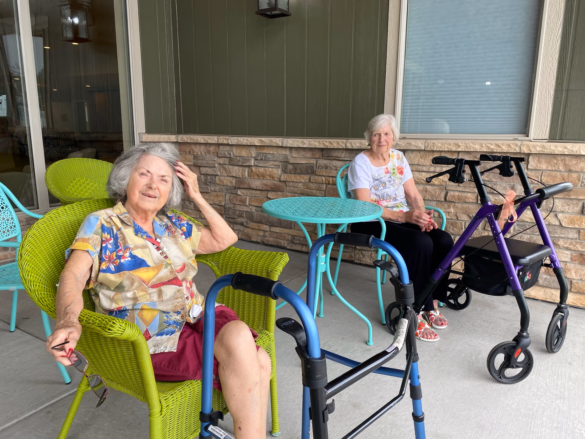 Two elderly women sitting outside on a patio area of an assisted living facility. One woman is seated in a bright green wicker chair wearing a colorful shirt and holding glasses, while the other woman is seated in a turquoise metal chair wearing a white floral shirt. There are two walkers in front of them and a turquoise metal table between them. The background shows a stone wall and a window with a closed blind.
