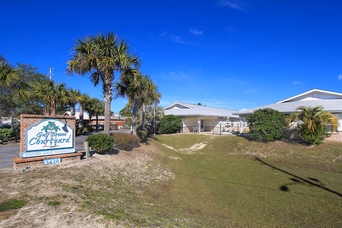 Exterior view of Gulf Breeze Courtyard Assisted Living with palm trees, an entrance sign, and single-story buildings under a clear blue sky.