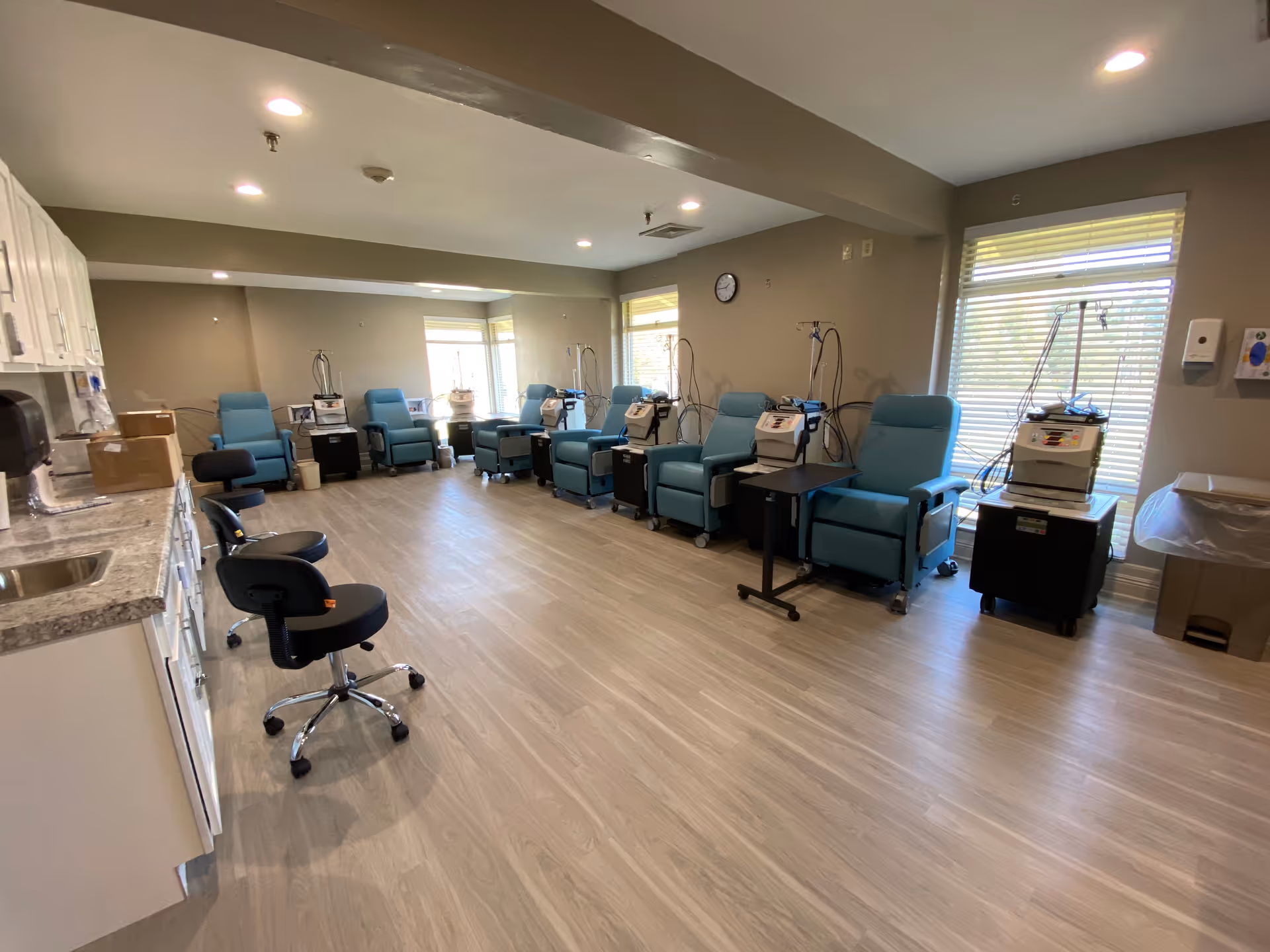 Bright clinical treatment room with teal reclining chairs arranged along the walls and dialysis/medical machines at each station.