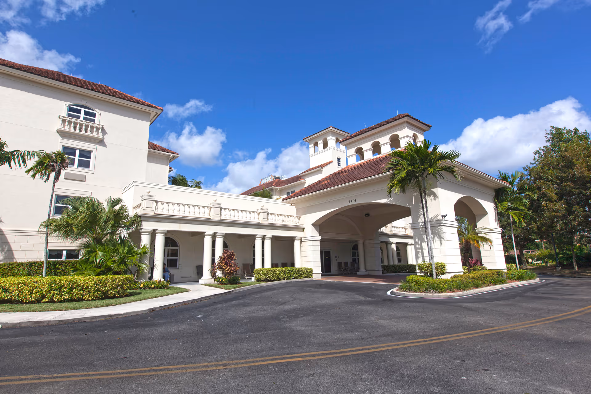 White, Mediterranean-style senior living building with arches, palm trees, and a curved driveway.