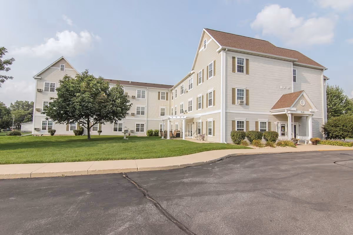 Three-story light-colored senior apartment building with a lawn, tree, and paved driveway under a partly cloudy sky.