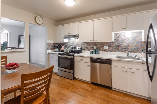 A modern kitchen with white cabinets, a stainless steel stove, dishwasher, and refrigerator. The backsplash features a colorful mosaic tile design. There is a wooden dining table with chairs and a bowl of red apples on the table. The floor is wooden, and a clock is mounted on the wall above a pass-through window to another room.