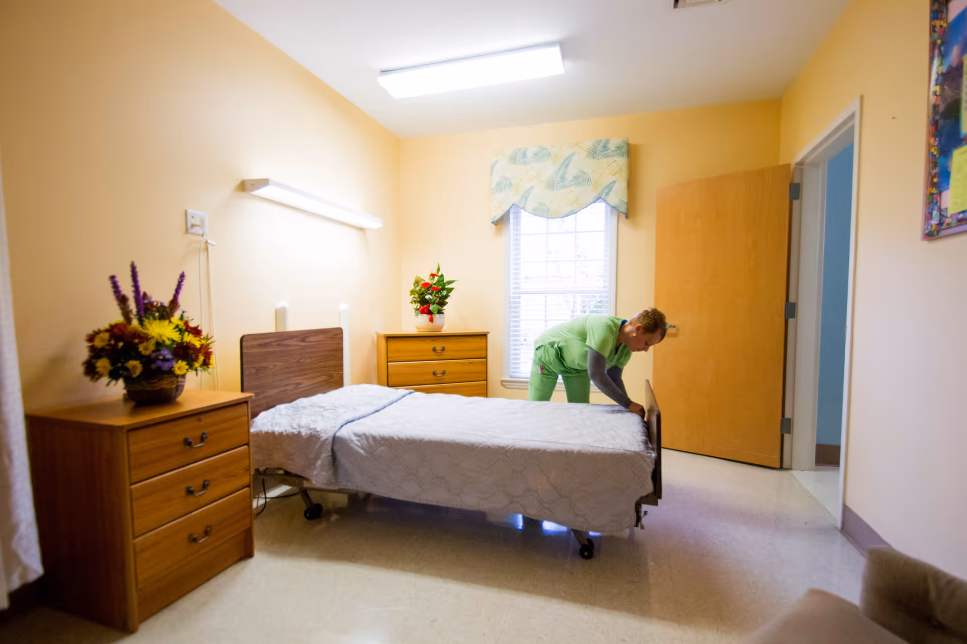 A caregiver in green scrubs is making a bed in a simple, well-lit bedroom with beige walls. The room contains a bed with a white quilt, two wooden dressers with flower arrangements on top, a window with a patterned valance, and an open door leading to another room.