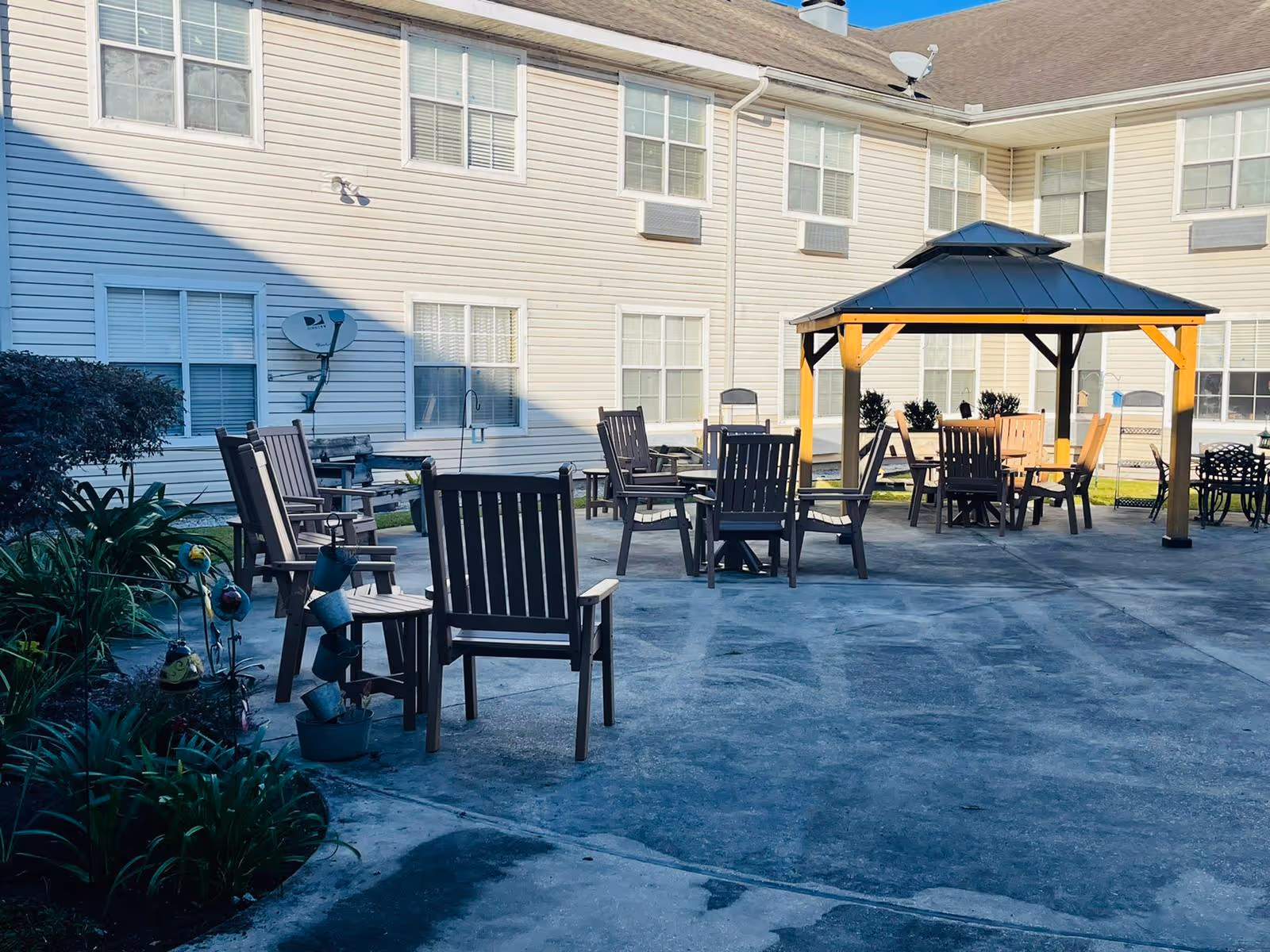 Outdoor patio area at Maison Jardin Living Community with multiple wooden chairs and tables arranged on a concrete surface. There is a gazebo with a dark roof providing shade over some seating. The background shows the exterior of a two-story building with multiple windows and air conditioning units.