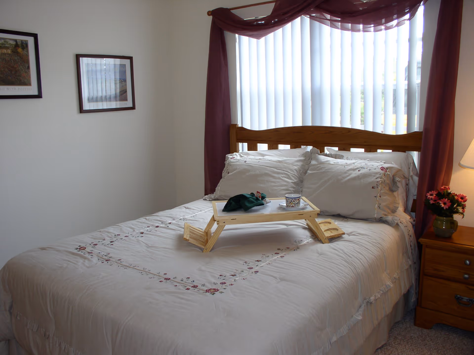 A neatly made bed with floral embroidered bedding and pillows, a wooden breakfast tray with a cup and napkin on top, positioned in front of a window with vertical blinds and purple curtains. A wooden nightstand with a lamp and a small vase of flowers is beside the bed, and two framed pictures hang on the adjacent wall.
