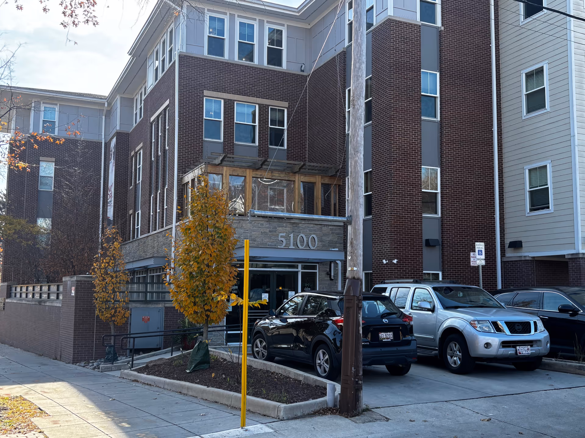 Exterior front entrance of a multi-story brick senior living building showing the address 5100, parked cars, and small trees.
