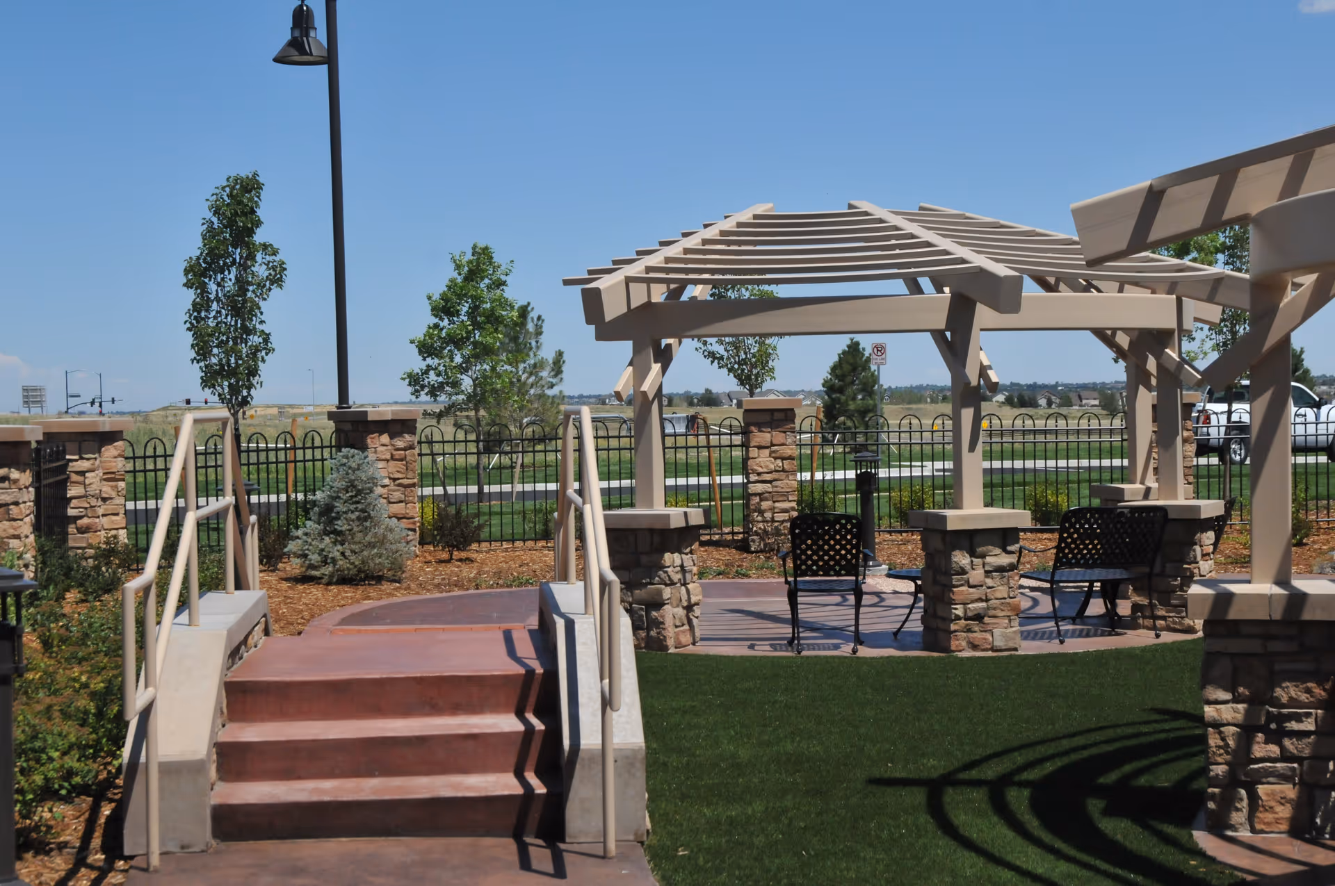 Outdoor seating area with a pergola structure supported by stone pillars, metal chairs, a small table, and a ramp with handrails leading up to the area. The background shows a fenced grassy area with trees and a clear blue sky.
