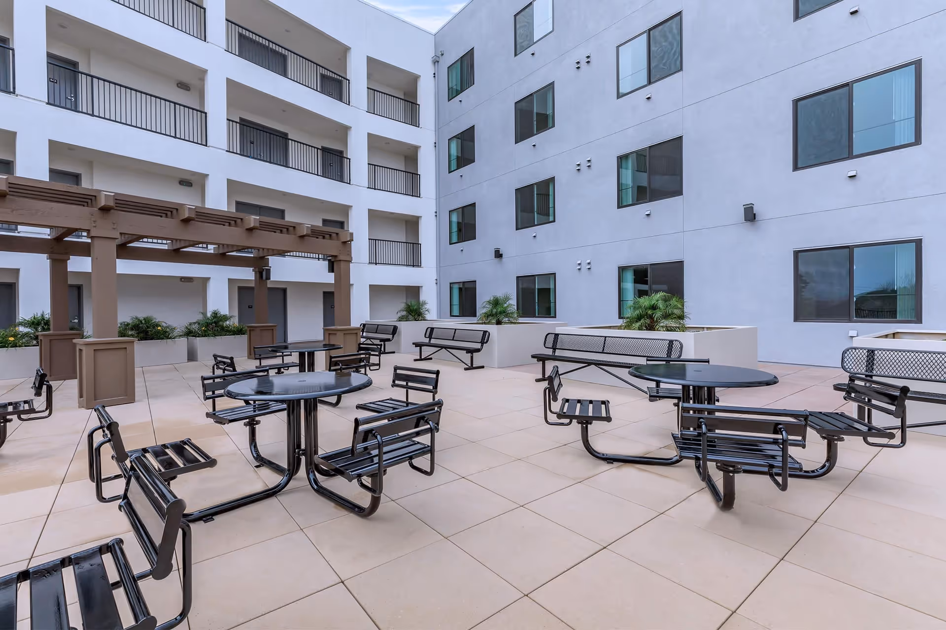 Open-air apartment courtyard with round metal picnic tables and chairs, a pergola, planters, and surrounding multi-story balconies.