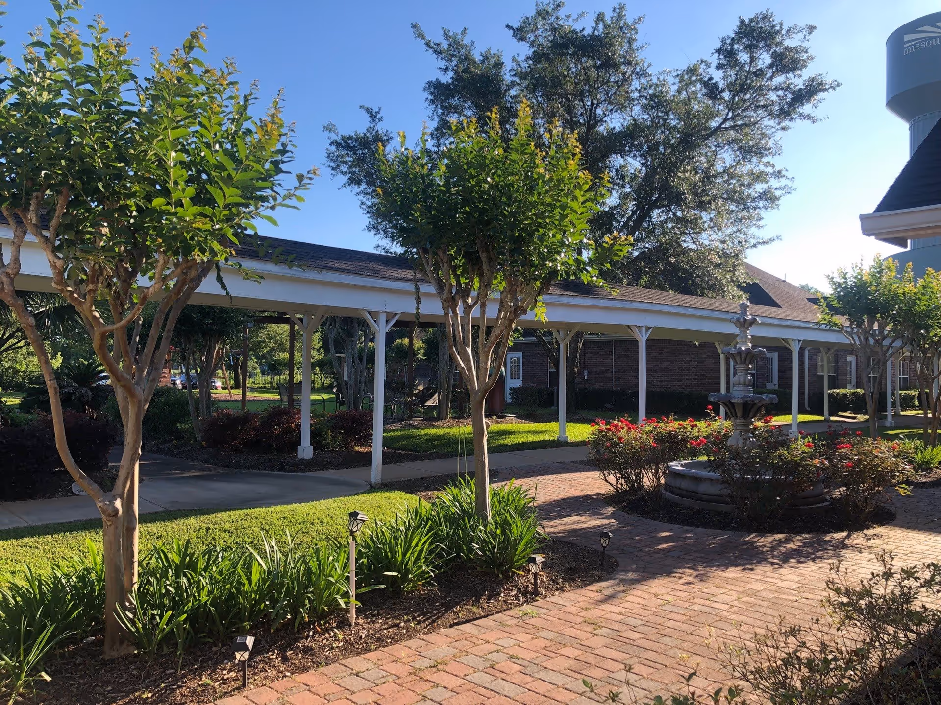 Sunny courtyard with a covered walkway, fountain, trees, and a brick path at a senior living facility.