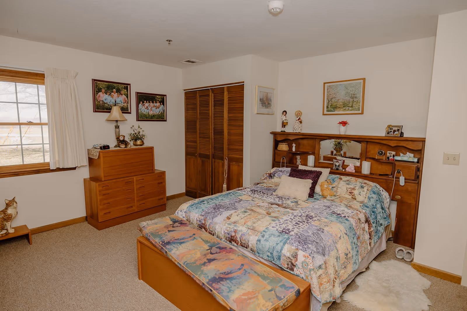 A cozy bedroom with a large bed covered in a colorful patchwork quilt, two pillows, and a small stuffed animal. The bed has a wooden headboard with shelves holding decorative items and a mirror. There is a wooden bench with a cushioned top at the foot of the bed. On the left side of the room, there is a wooden dresser with a lamp, a teddy bear, and a flower arrangement. Two framed family photos hang on the wall above the dresser. A window with white curtains lets in natural light. The room has beige carpet and white walls with additional framed artwork and a small closet with wooden louvered doors.