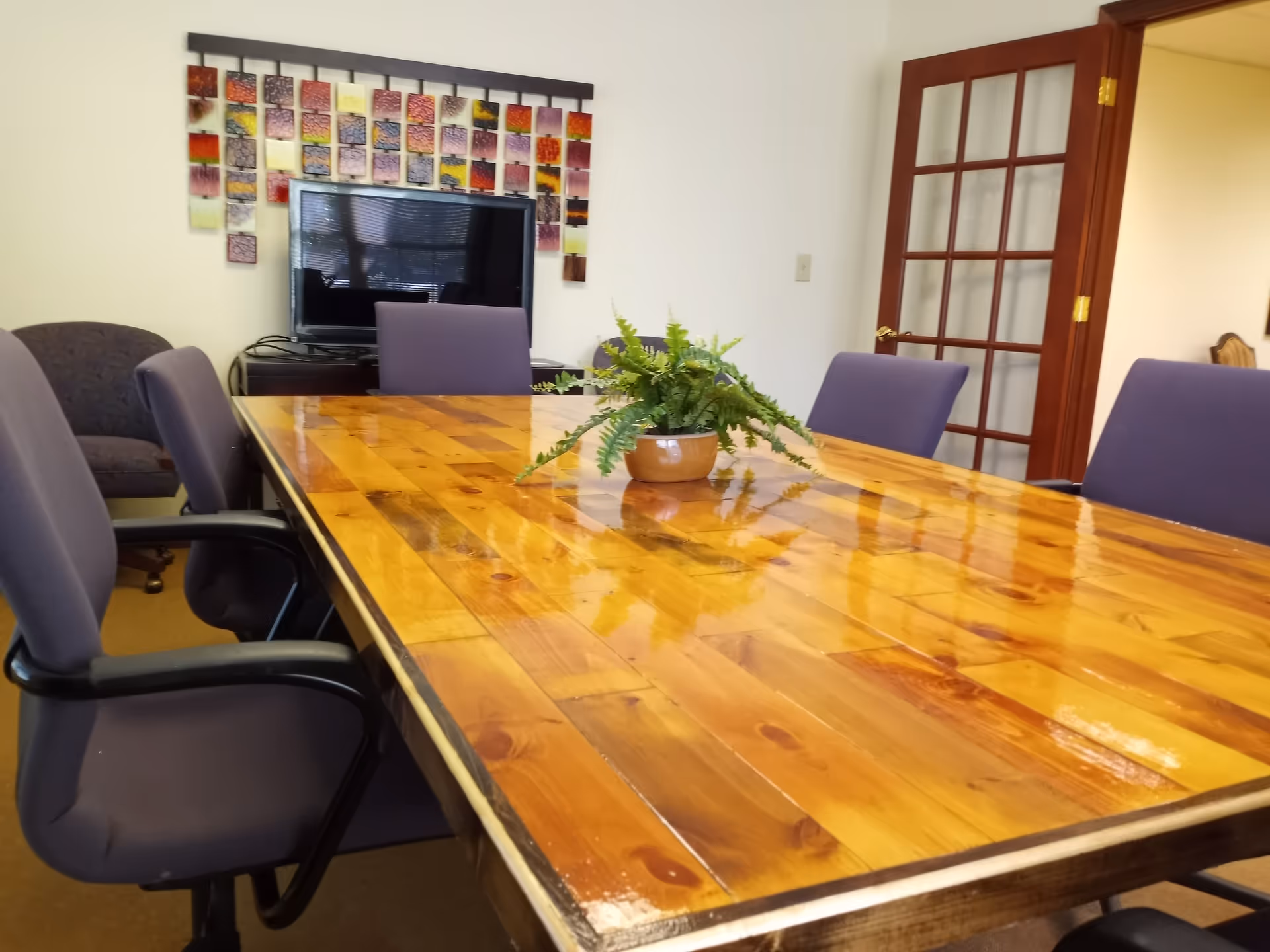 A long wooden conference table with purple chairs, fern centerpiece, and art in a meeting room.