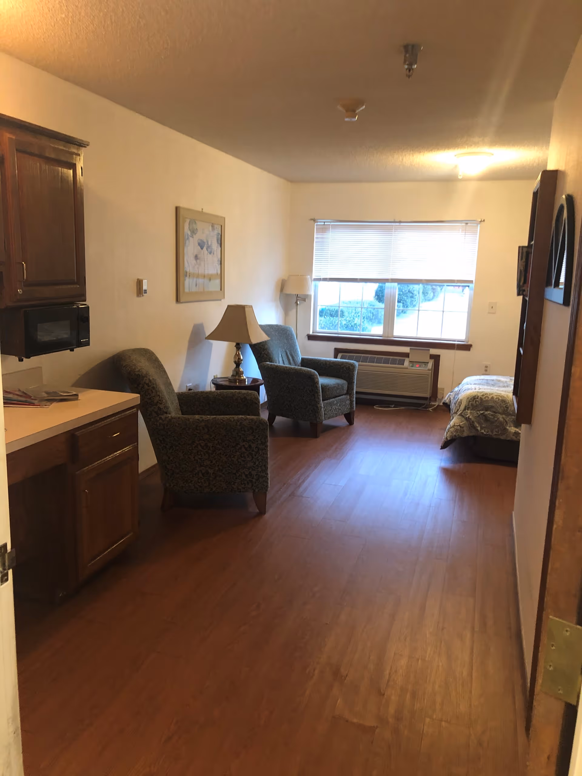 Interior view of a senior living facility room with wooden flooring, two upholstered armchairs, a side table with a lamp, a bed near a window with blinds, a wall-mounted microwave, and wooden cabinets.