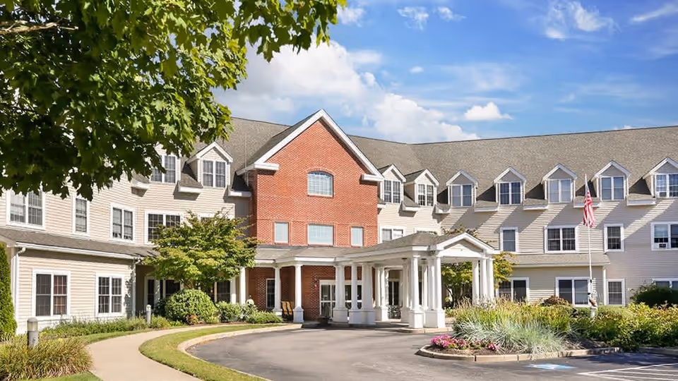Front exterior of a multi-story senior living building with a covered porte-cochère entrance, landscaped driveway, and an American flag.