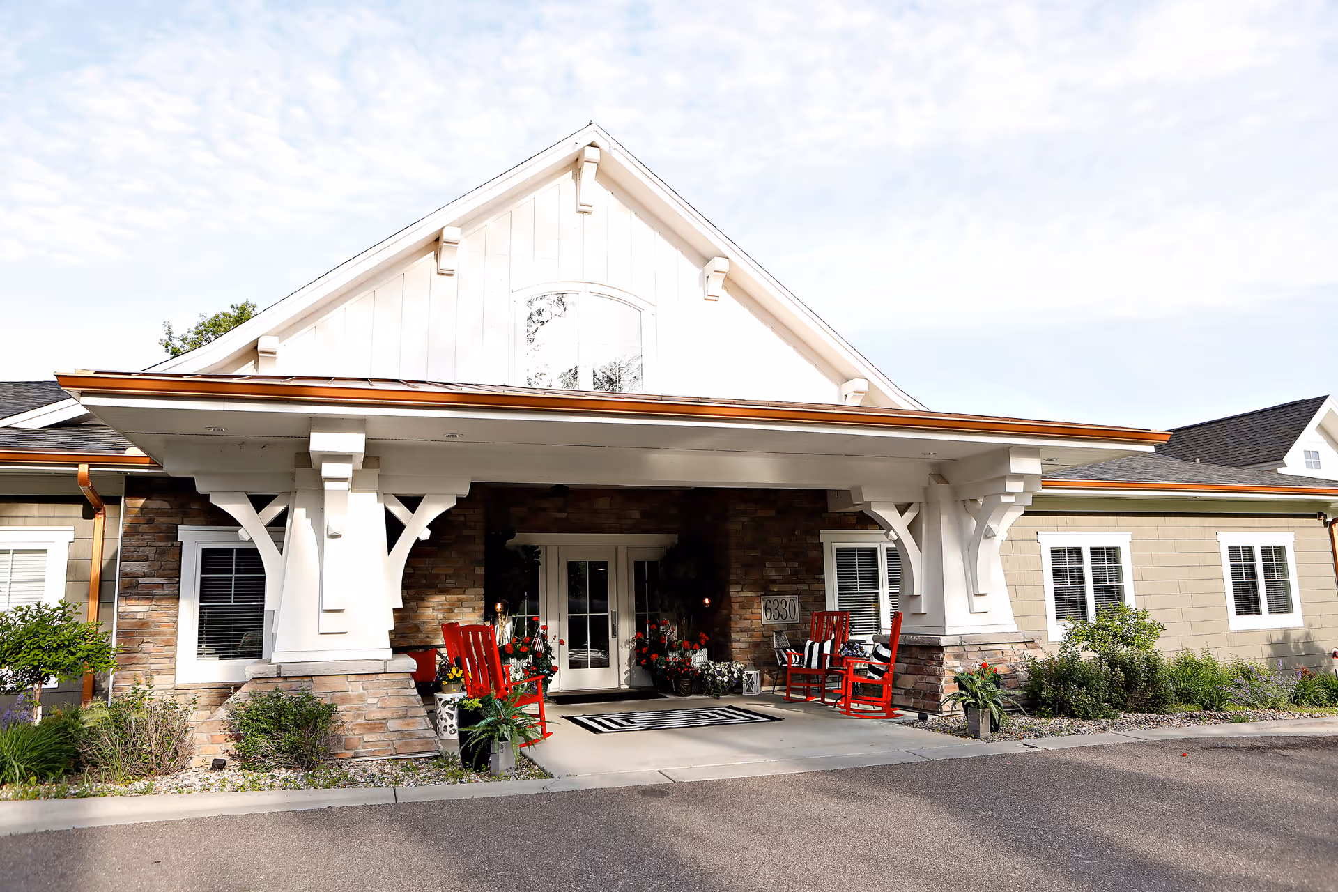 Covered front entrance of a memory care facility with a gabled roof, red rocking chairs, potted plants, and double doors.