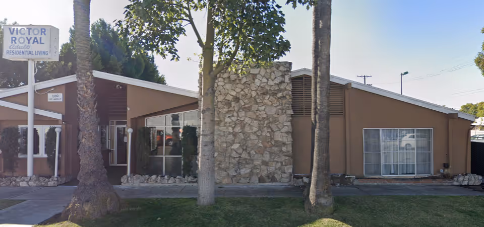 Exterior view of Victor Royale Adult Residential Living facility showing a single-story building with a stone chimney, large windows, and a sign with the facility name. There are palm trees and a sidewalk in front of the building.