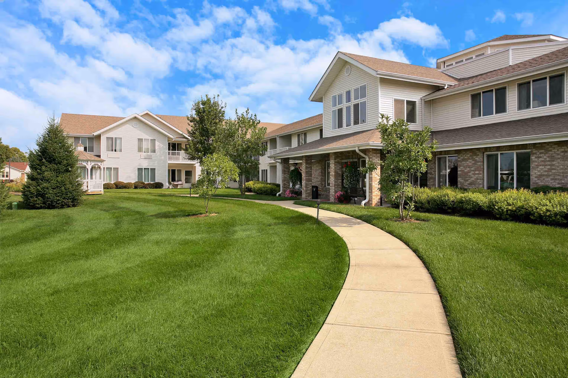 Two-story senior living building with a covered entrance, curved sidewalk, and manicured lawn under a blue sky.