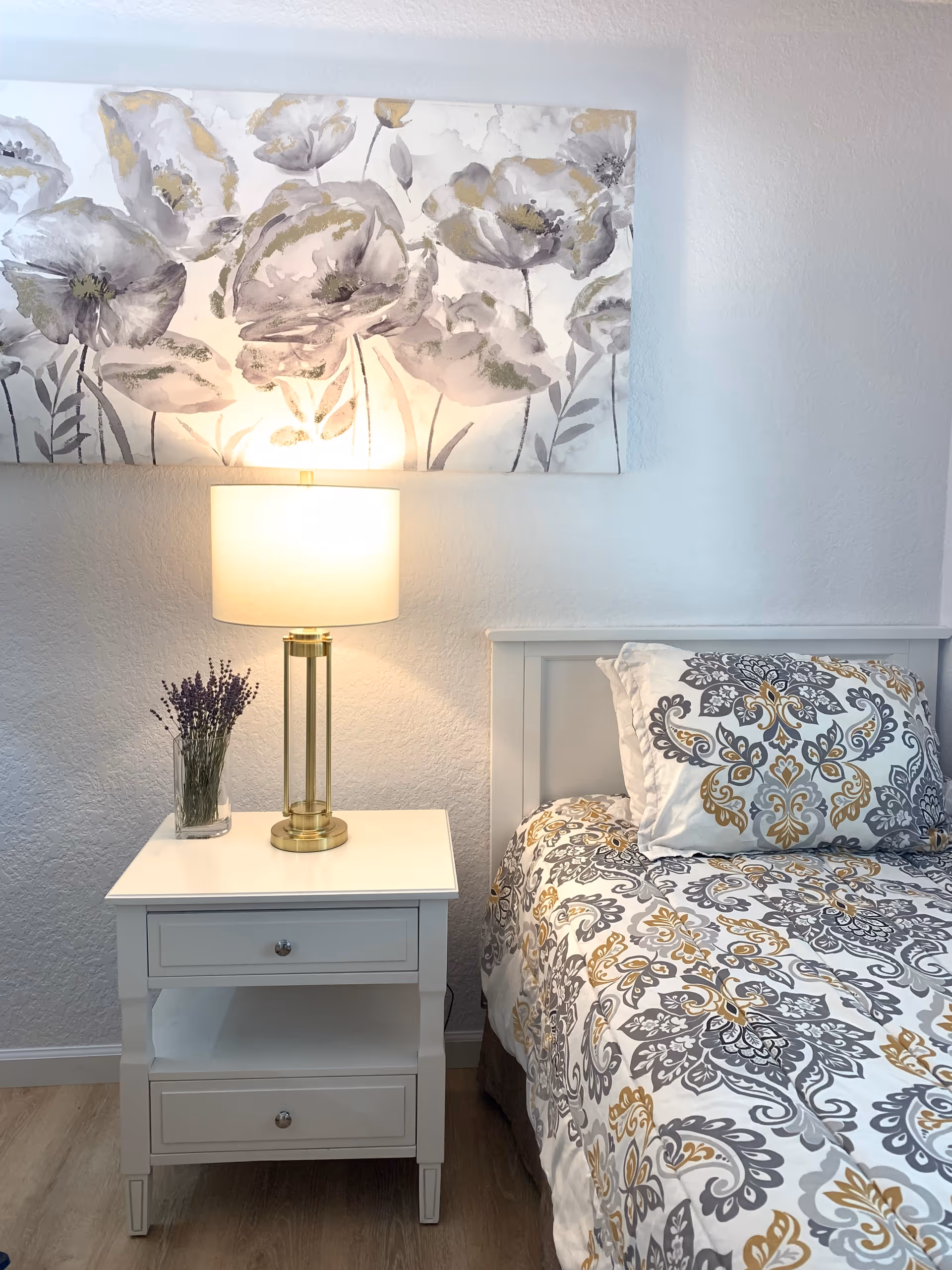 A cozy bedroom corner featuring a white nightstand with two drawers, a brass and glass table lamp with a white shade, and a small vase of lavender. Next to the nightstand is a bed with a white headboard and bedding with a gray, gold, and white floral pattern. Above the nightstand hangs a large floral painting in muted tones.