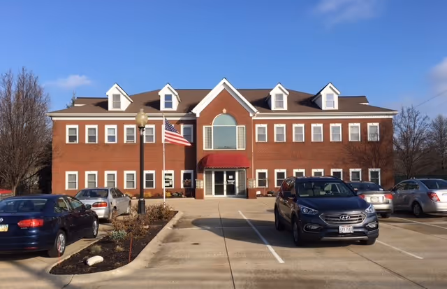 Front view of a brick senior living facility with parked cars and American flag.