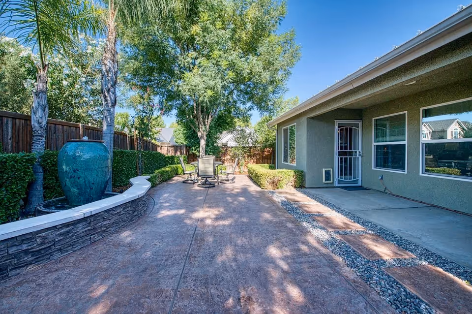 Outdoor patio area with a stone water fountain on the left, surrounded by greenery and palm trees. There is a paved walkway leading to a seating area with a table and chairs under a large tree. On the right side, there is a building with windows and a door with a security screen.
