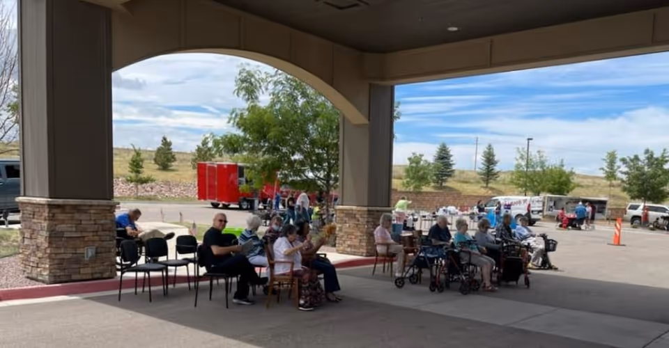 Residents seated under a covered building entrance with walkers and chairs while vehicles and trailers are parked in the driveway.