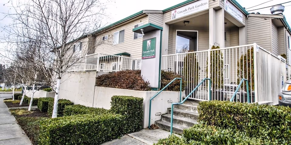 Exterior view of Cascade Park Gardens facility showing a two-story beige building with white railings, stairs leading up to the entrance, trimmed bushes, and leafless trees along the sidewalk.