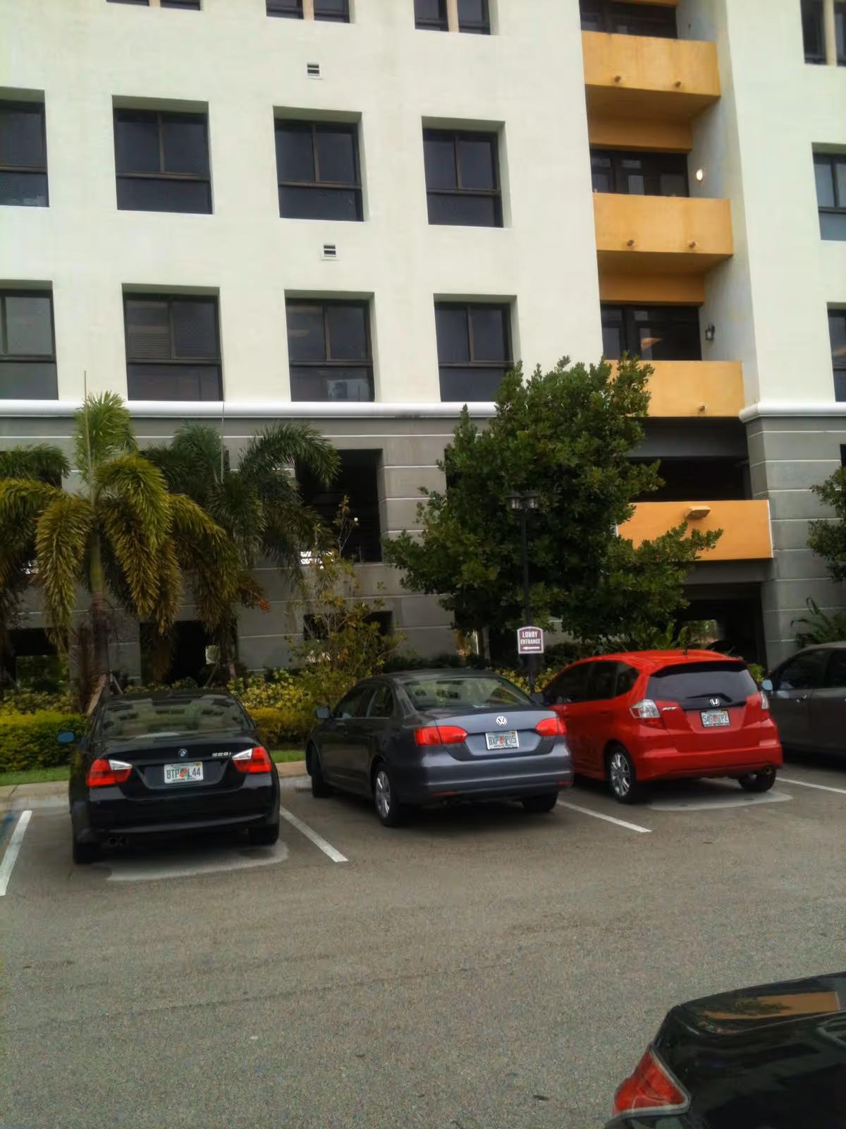 Modern multi-story building with orange balconies, palm trees, and a parking lot.