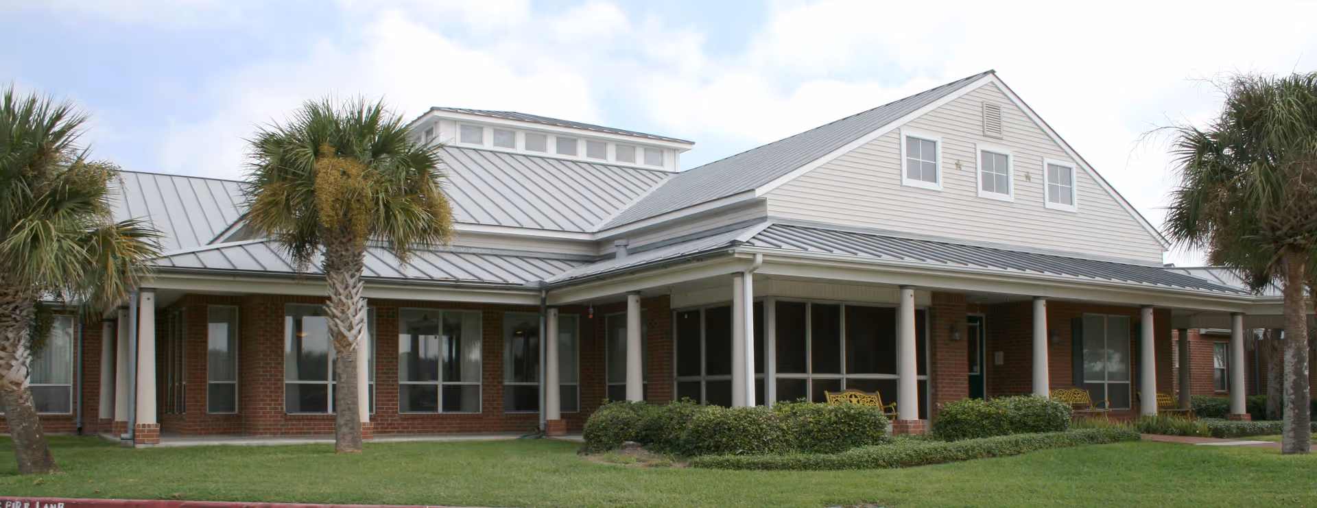 Front exterior of a single-story assisted living building with a covered porch, columns, palm trees and a manicured lawn.