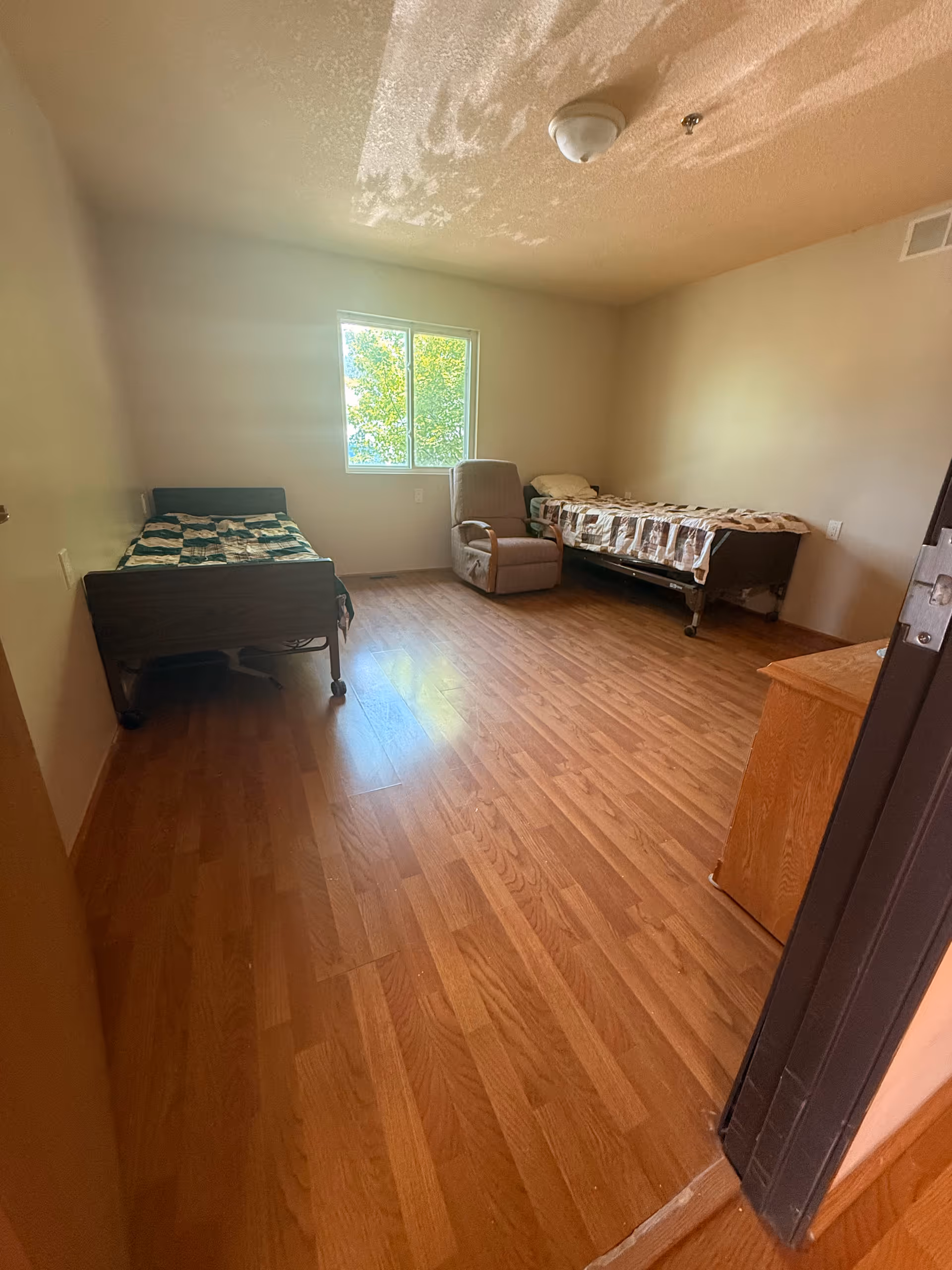 Small bedroom in a senior facility with two beds, a chair, and a window.