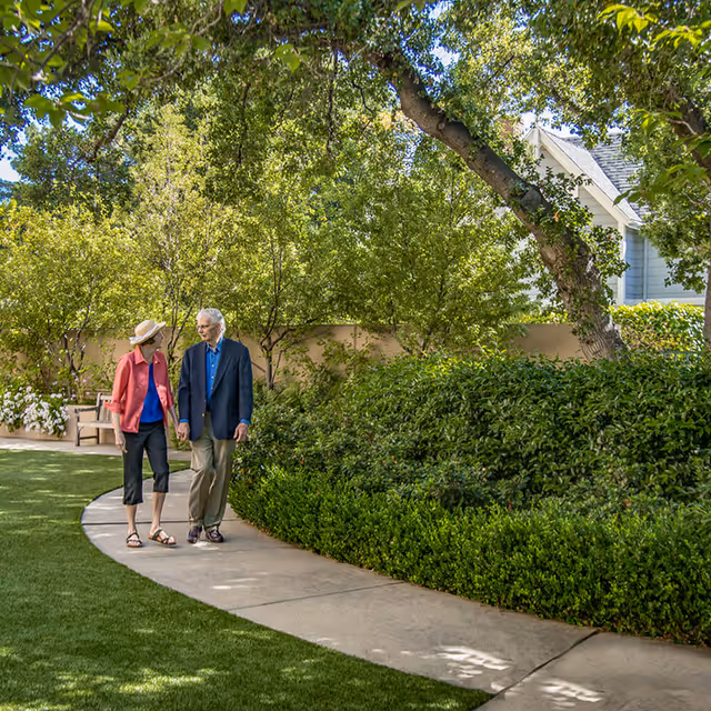 An elderly couple walking hand in hand along a curved garden pathway surrounded by lush green bushes and trees, with a bench and a house partially visible in the background.