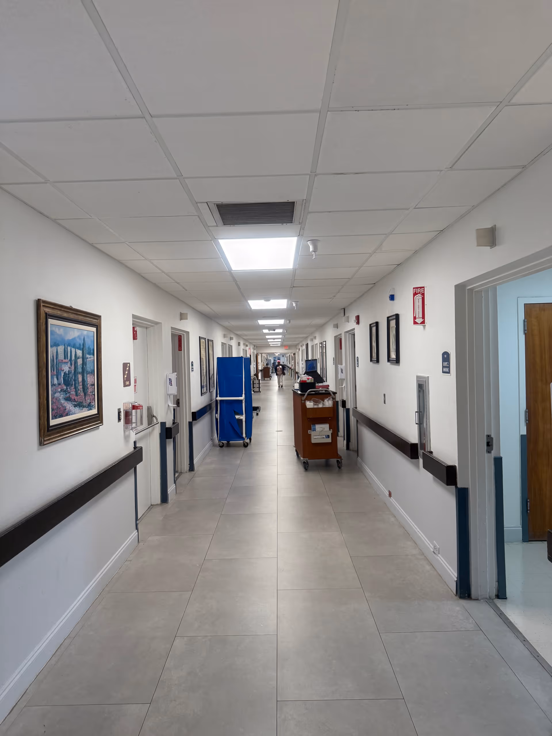 Long, well-lit hospital-style corridor inside a senior living facility with carts and framed artworks.