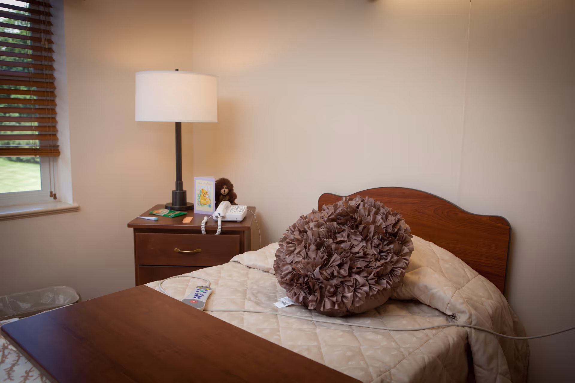 A cozy bedroom with a wooden headboard bed covered with a beige quilt and a large decorative brown pillow. Next to the bed is a wooden nightstand with a white lamp, a telephone, a greeting card, and a small teddy bear. A window with wooden blinds is partially visible on the left side.