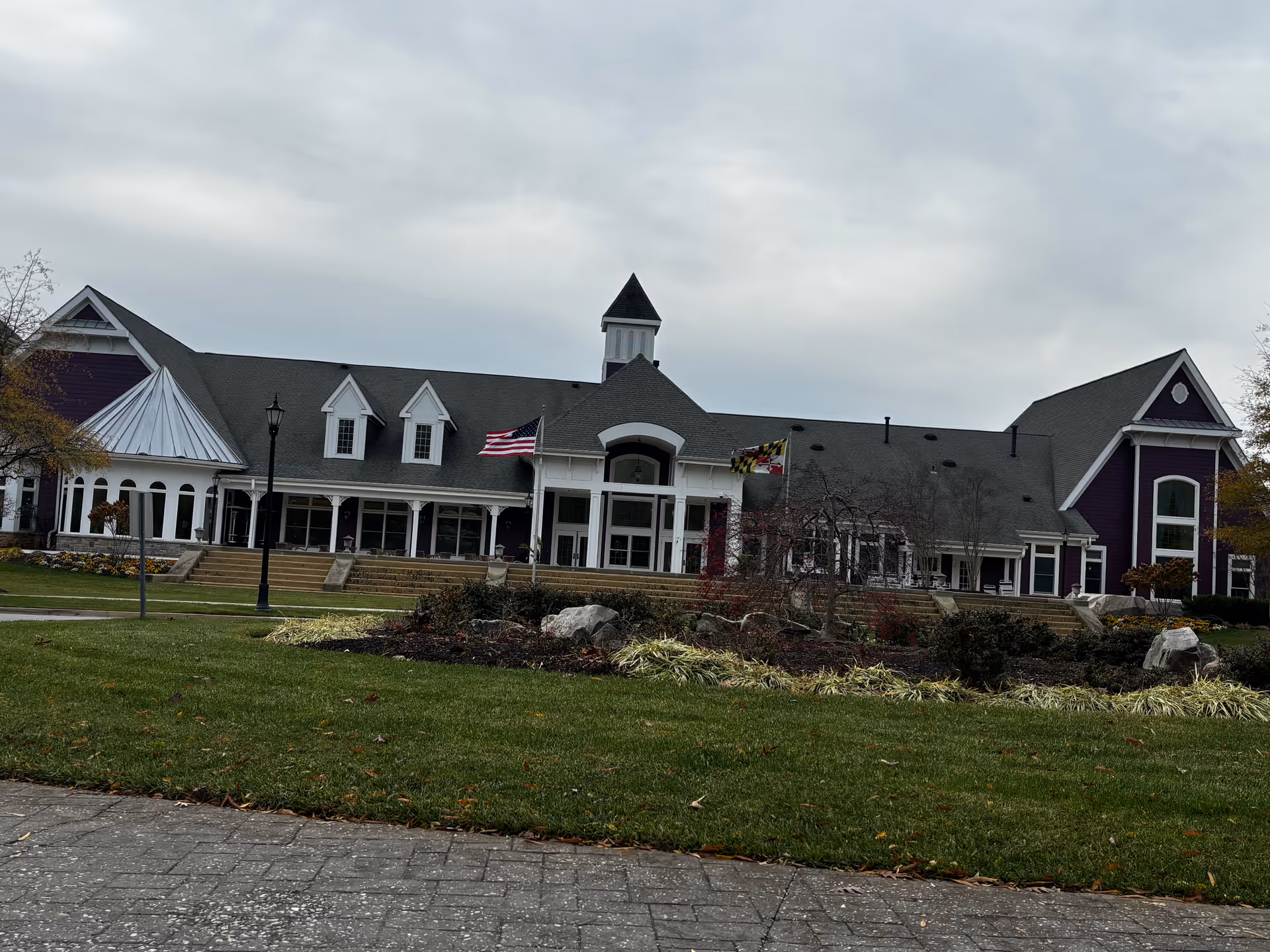 Exterior view of a large senior living facility building with a gray roof and purple siding. The building has multiple windows, dormers, and a central entrance with steps leading up to it. Two flags are flying near the entrance, and there is a landscaped garden with shrubs and rocks in front of the building. The sky is overcast.