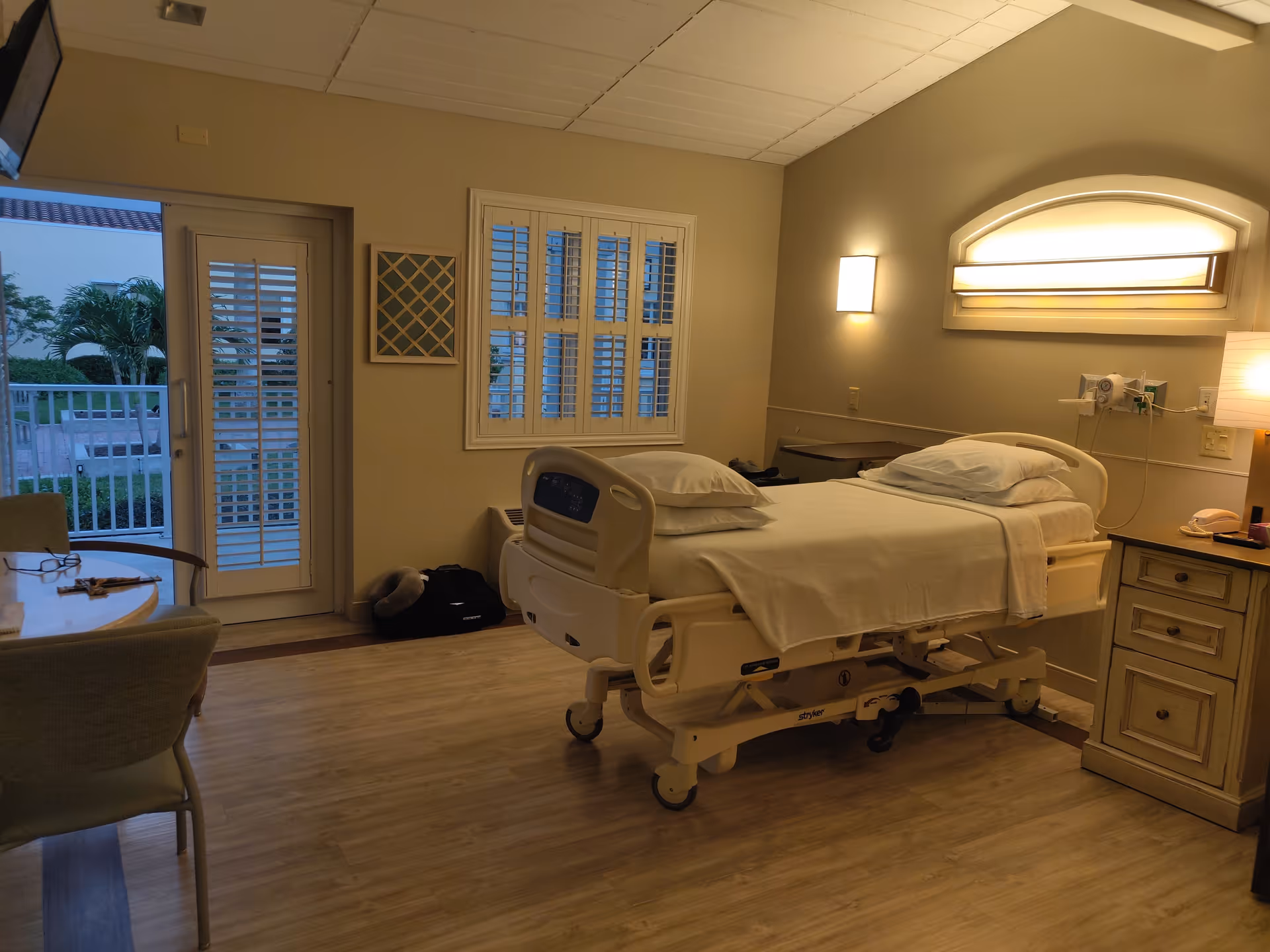 Senior living patient room with adjustable bed, window, balcony door, and bedside furniture.