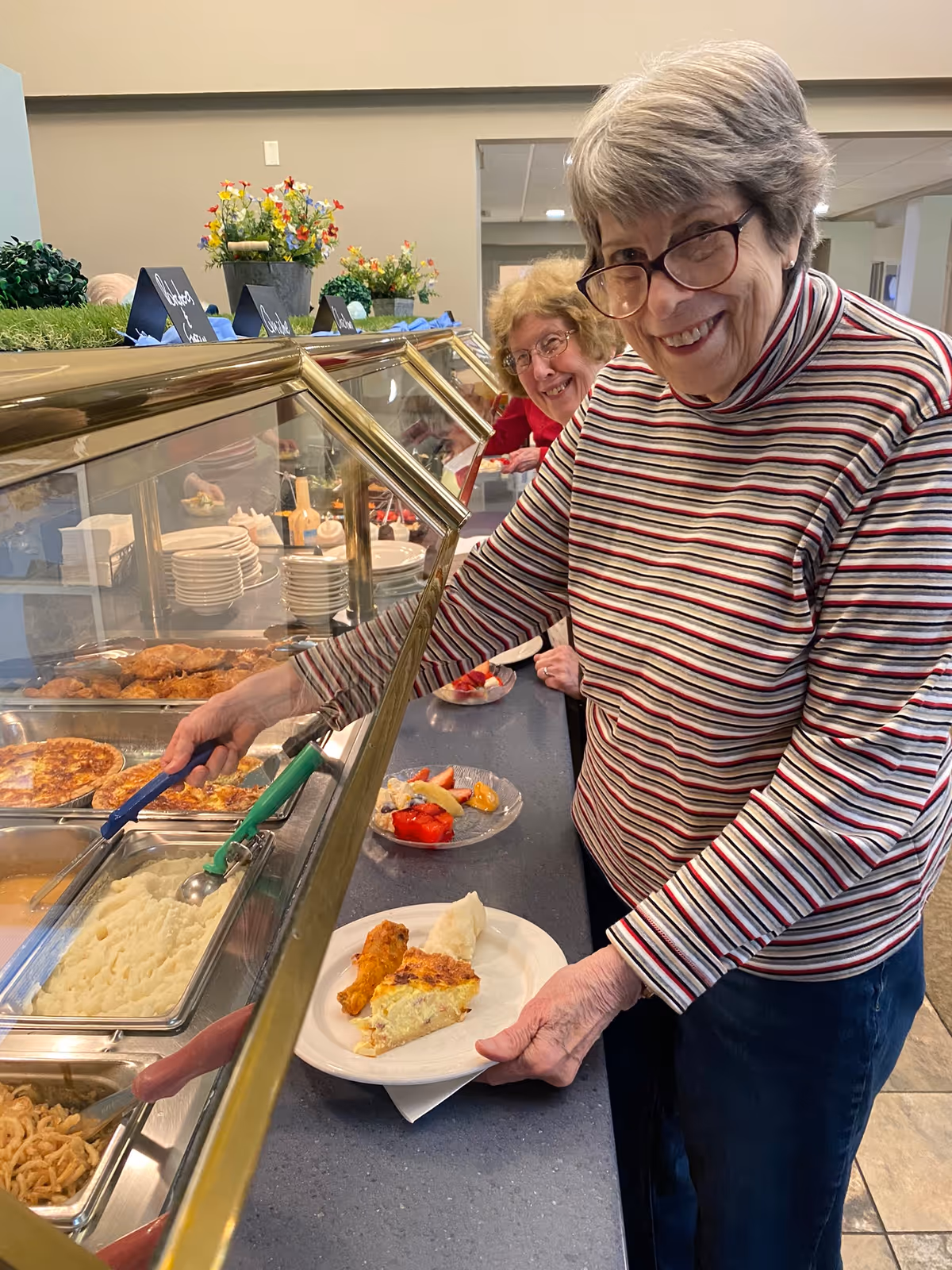 Two elderly women smiling as they serve themselves from a buffet line in a dining room.