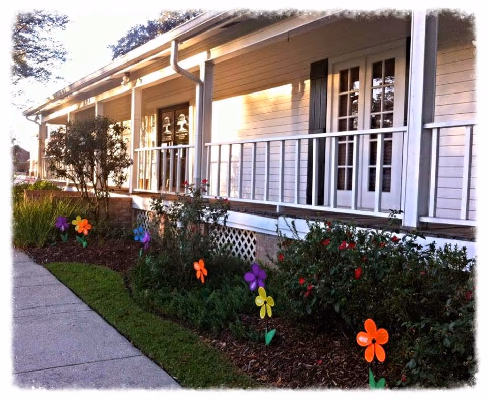 A single-story senior facility with a covered porch, railing, and colorful flower garden.